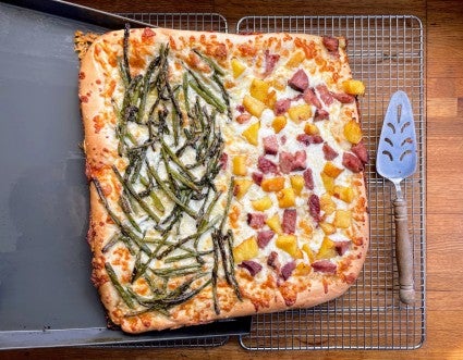 Baked square pizza being slid out of its pan onto a cooling rack.